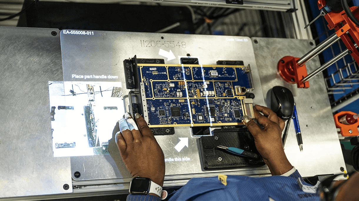 Blick über die Schulter auf einen Mitarbeiter bei L3Harris, der Elektronik mithilfe von LightGuide projizierten Augmented-Reality-Arbeitsanweisungen montiert